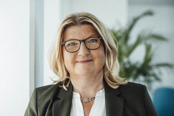 A portrait photograph of a woman wearing glasses, a white blouse and an olive green blazer.