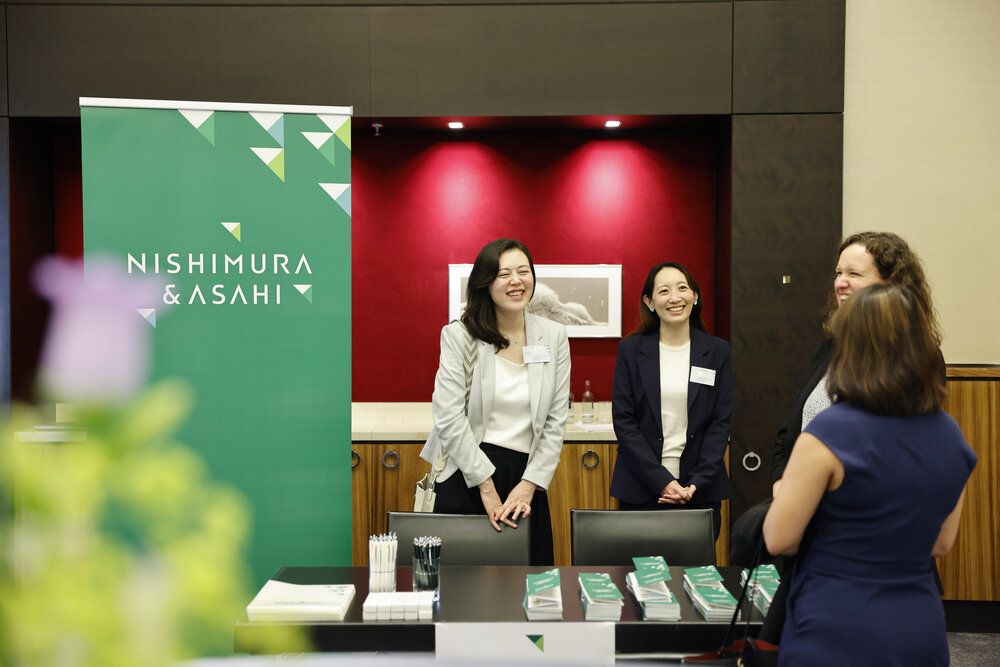 Four women are chatting at a trade fair stand, a table with brochures and pens, behind them a green banner with the words Nishimura & Asahi.