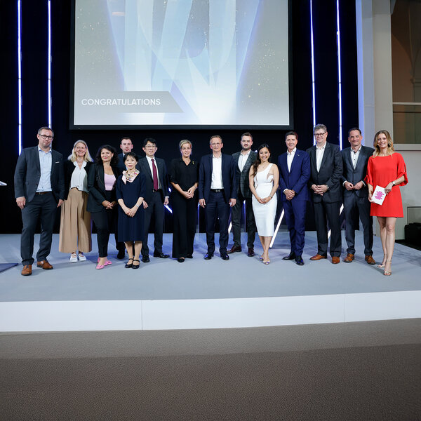Fourteen people in business attire stand in a group, with the word “Congratulations” displayed on a large monitor behind them.