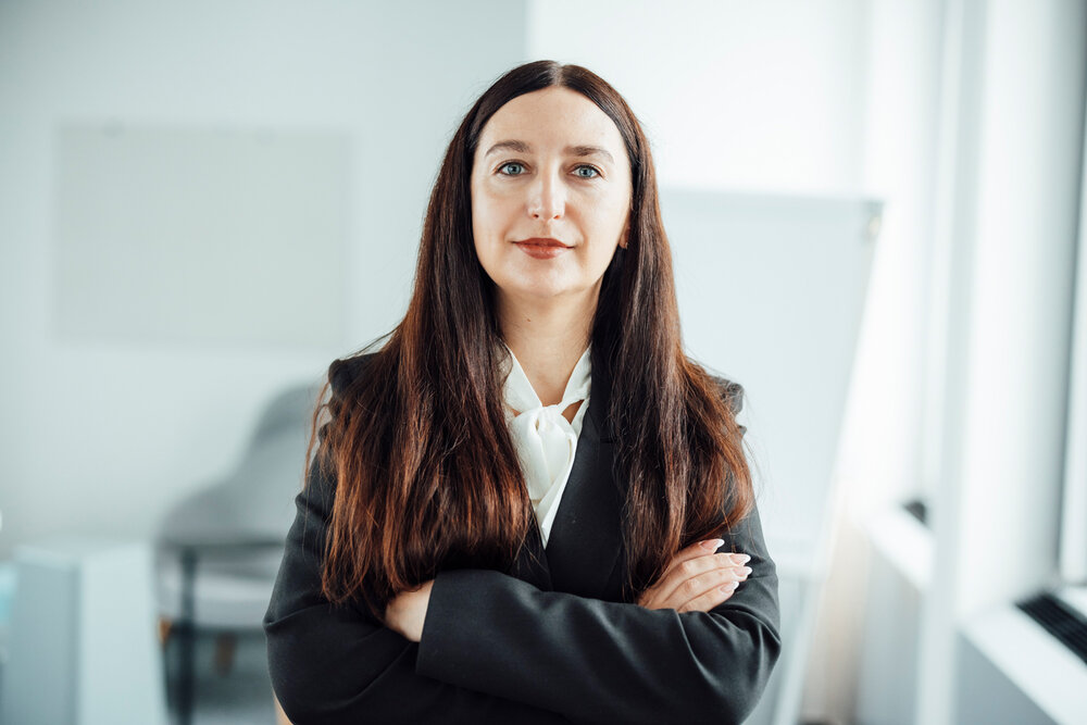 A portrait photograph of a smartly dressed woman wearing a white blouse and black blazer, with her arms crossed.