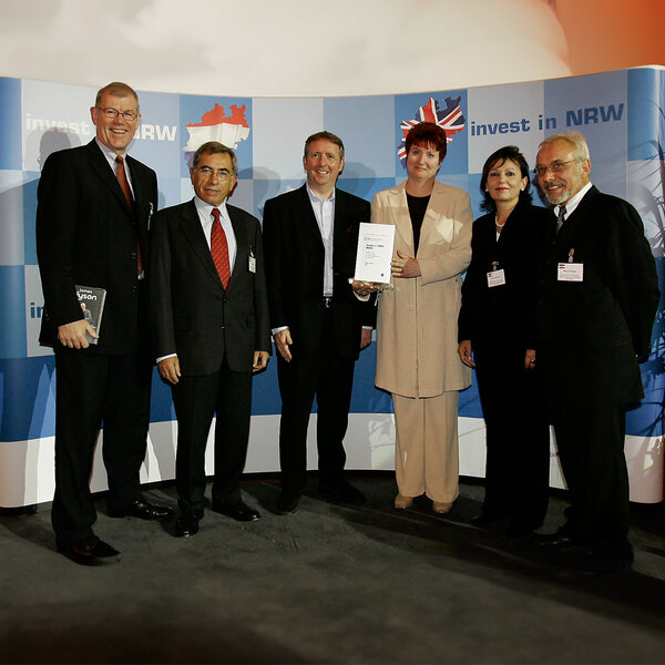 Six people are standing in suits and business attire in front of a wall with the words ‘invest in NRW’ written on it, and a woman is holding a certificate.