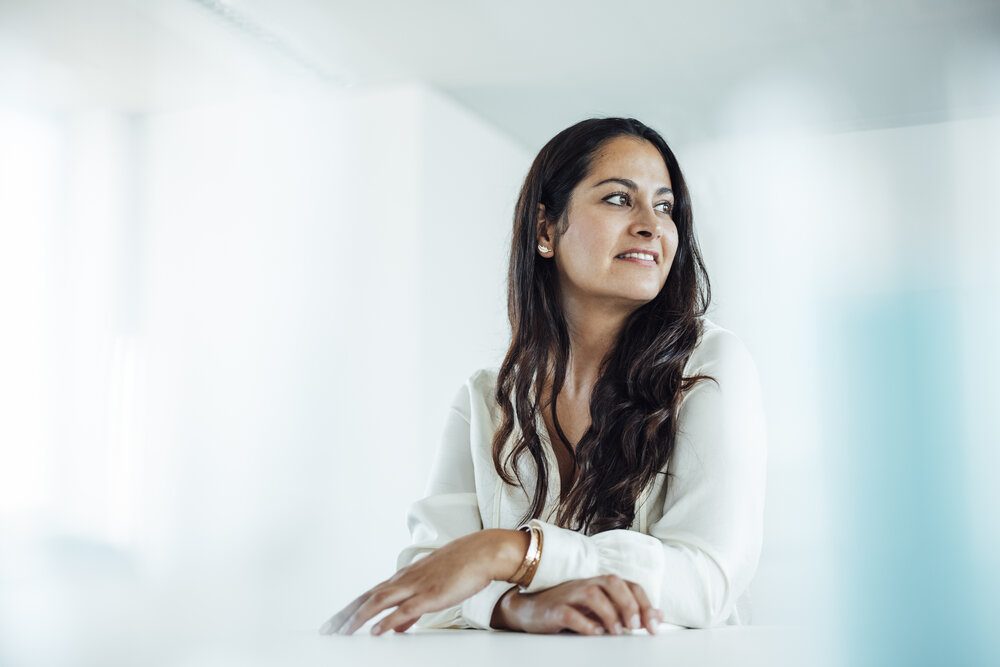 A portrait photograph of a young woman sitting behind an office desk with her arms resting on it. She is looking to the side.