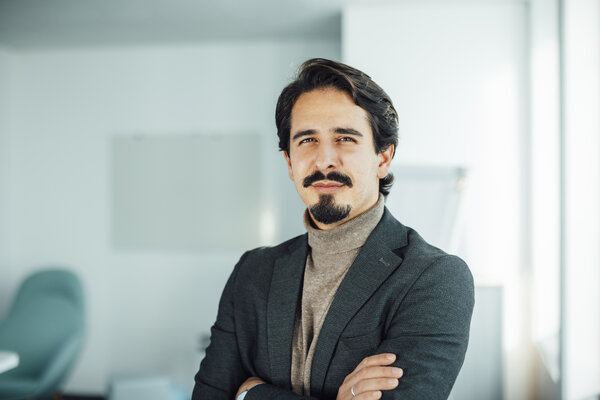 A portrait photo of a man wearing a brown sweatshirt with a shawl collar and a black and grey jacket. His arms are crossed