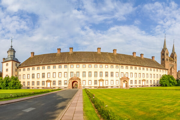 Eine Panoramaaufnahme von der Fassade des Klosters Corvey. Im Vordergrund sind große Rasenflächen und ein asphaltierter Weg zum Eingang.