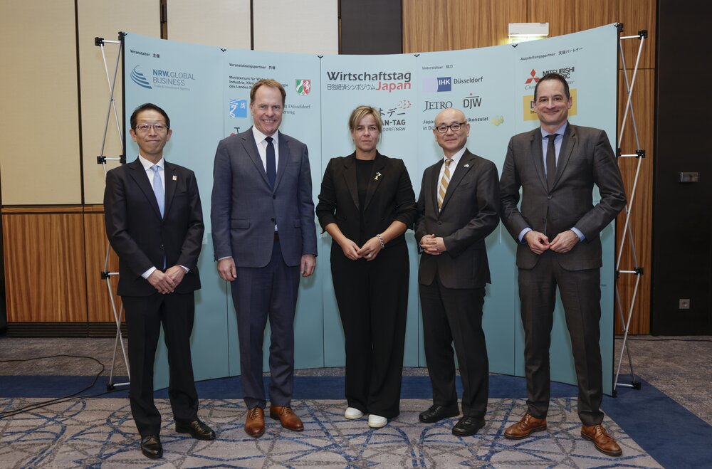 Five people in business attire stand in front of a wall with logos and lettering.