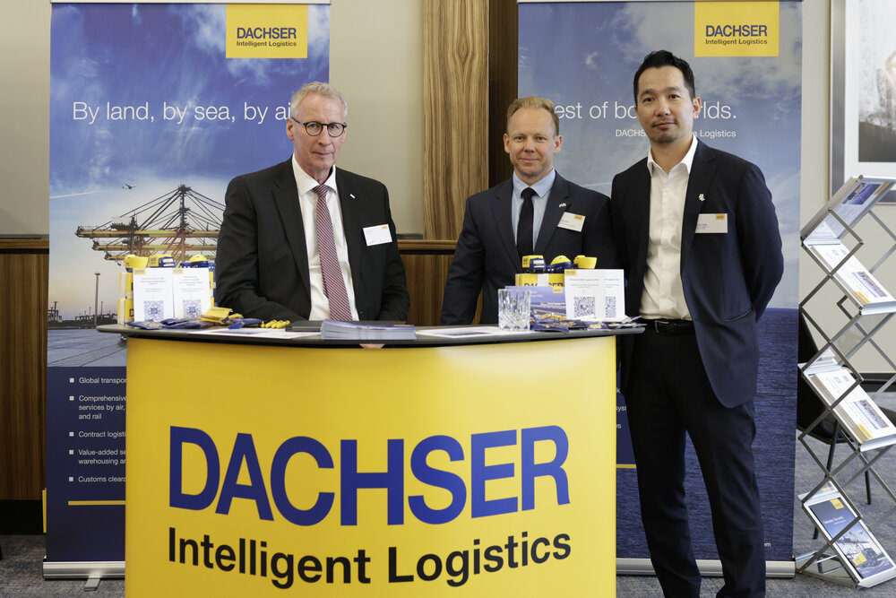 Three men in suits stand behind a yellow exhibition stand bearing the words “Dachser Intelligent Logistics”, with banners displayed in the background.