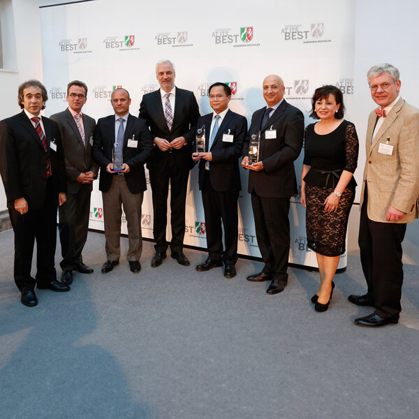 Nine people in business attire stand side by side, three holding glass trophies, with a screen in the background displaying the words “Germany at its best”.