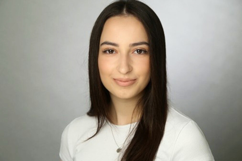 A portrait photograph of a young woman. She is wearing a light-coloured top and has dark hair.