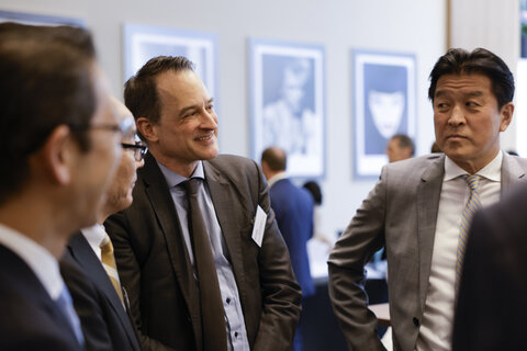 Several men in suits stand facing each other, two in the foreground of the photo, with a blurred background featuring framed pictures on the wall.