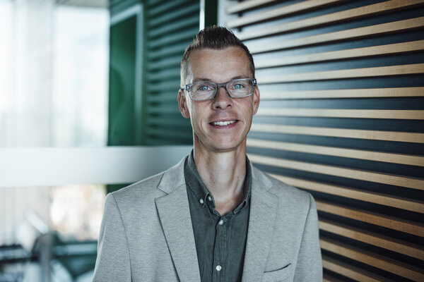 A portrait photograph of a man with short brown styled hair standing in front of a glass partition. He is wearing glasses.