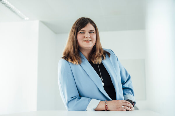 A portrait photograph of a young woman resting one arm on an office desk. She is wearing a light blue blazer and a black blouse.