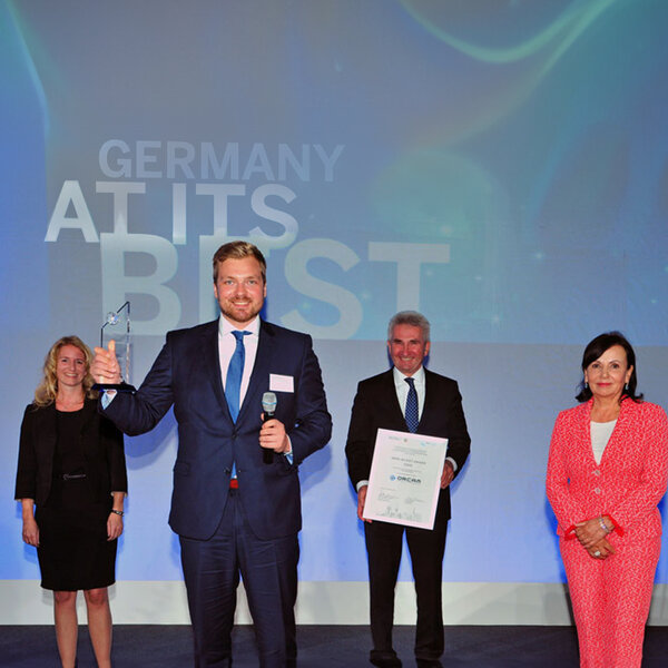 Four people in business attire are standing on a stage, one man is holding a glass trophy, another a certificate.