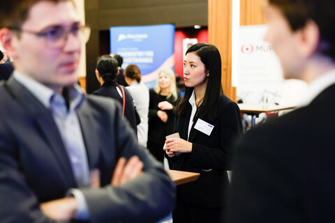 A woman in business attire with a name tag standing among several people, a blurred conversation in the foreground.