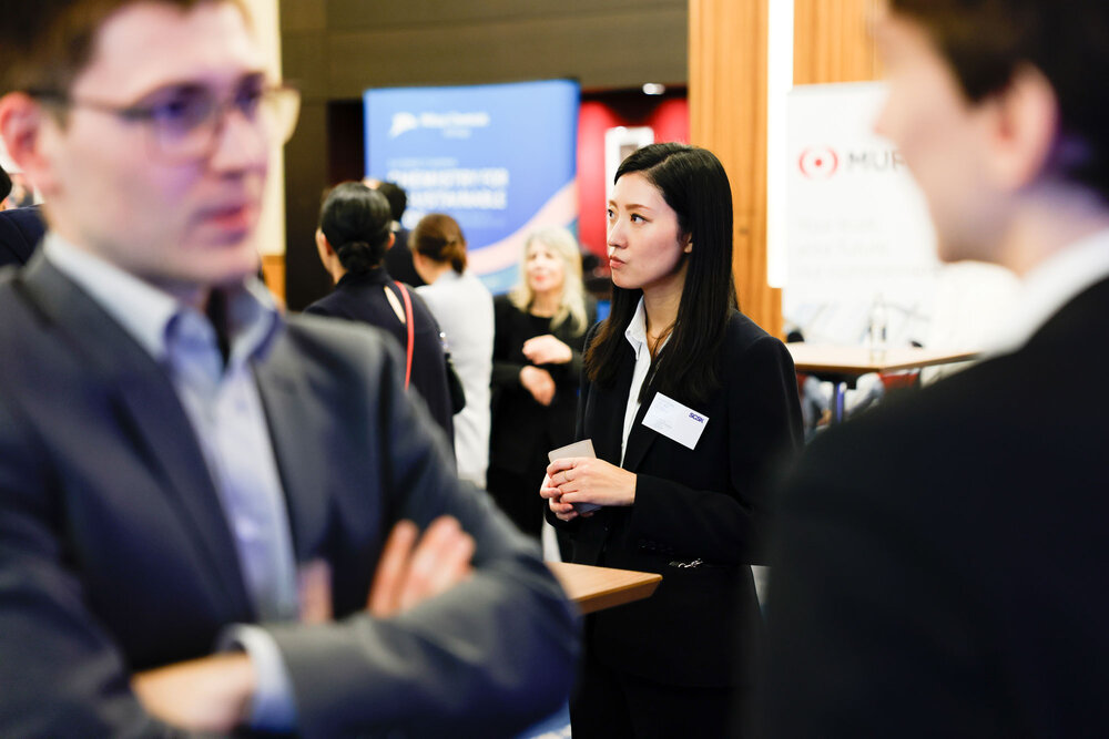 A woman in business attire with a name tag standing among several people, a blurred conversation in the foreground.