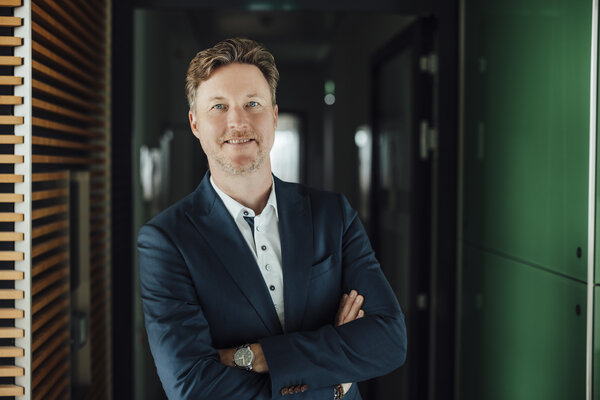A portrait photograph of a smartly dressed man with his arms crossed and leaning to one side in an office corridor.