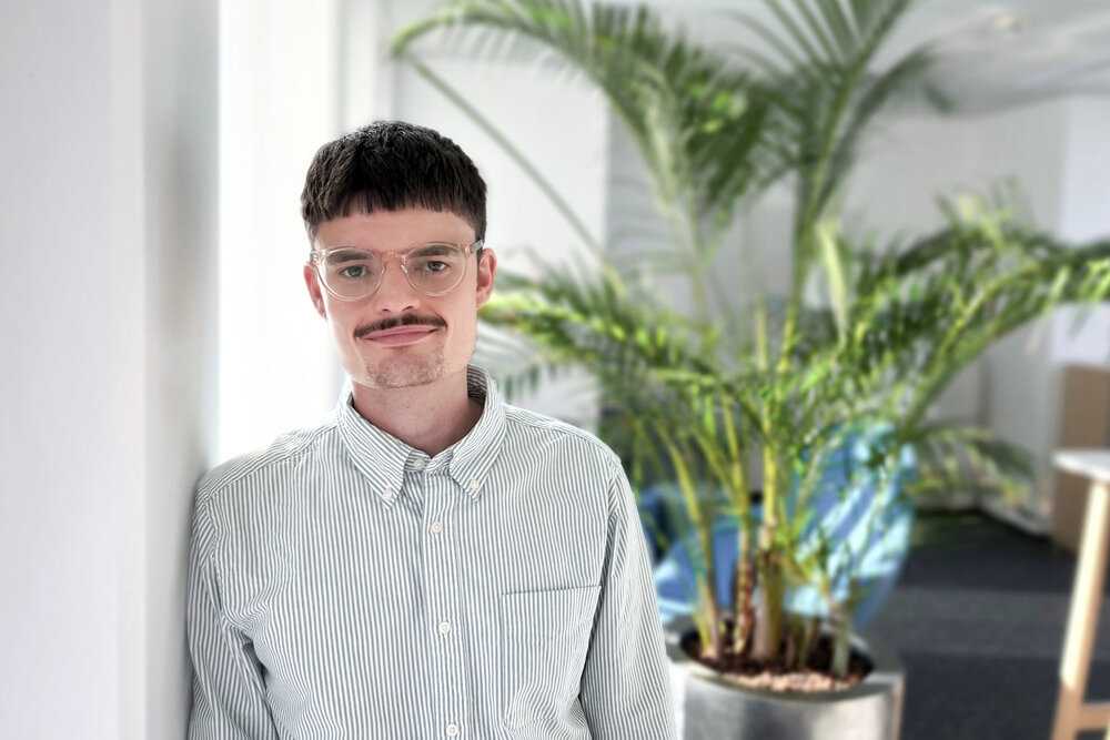 A portrait photo of a young man wearing glasses and a striped light-coloured shirt. He is leaning against an office wall.
