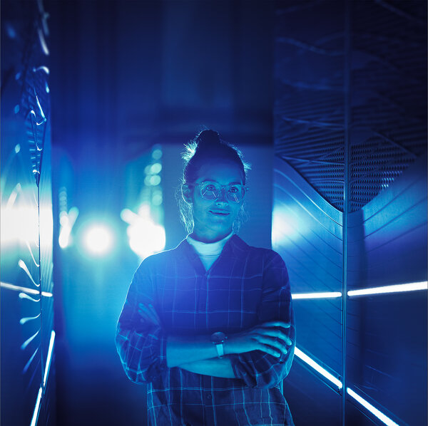 A young woman wearing glasses stands with her arms crossed in a blue-lit server room.