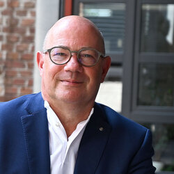 Ein Mann mit Brille, blauem Anzug und weißem Hemd lehnt entspannt an einer Armlehne und lächelt in die Kamera.