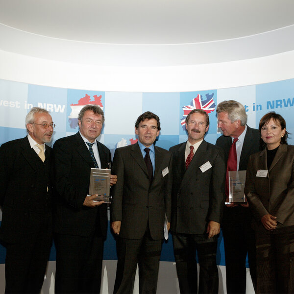 Six people in suits stand side by side in front of a wall with the words ‘Invest in NRW’ written on it, two of them holding awards in their hands.