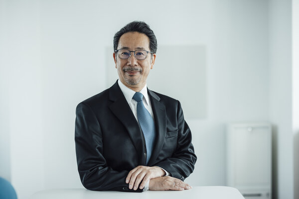 A portrait photograph of a smartly dressed man wearing glasses and sitting behind an office desk.
