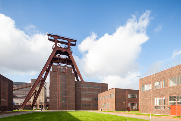 Das Industrie-Denkmal mit rotem Förderturm und Backsteingebäuden der Zeche Zollverein bei blauem Himmel mit weißen Wolken.