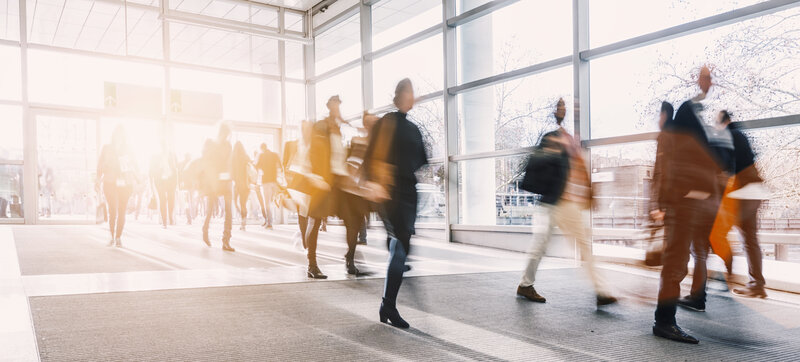 Blurred photograph of several people in motion as they enter or leave a light-filled hall with a glass front.