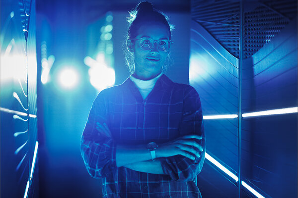 Eine junge Frau mit Brille steht mit verschränkten Armen in einem blau beleuchteten Serverraum.