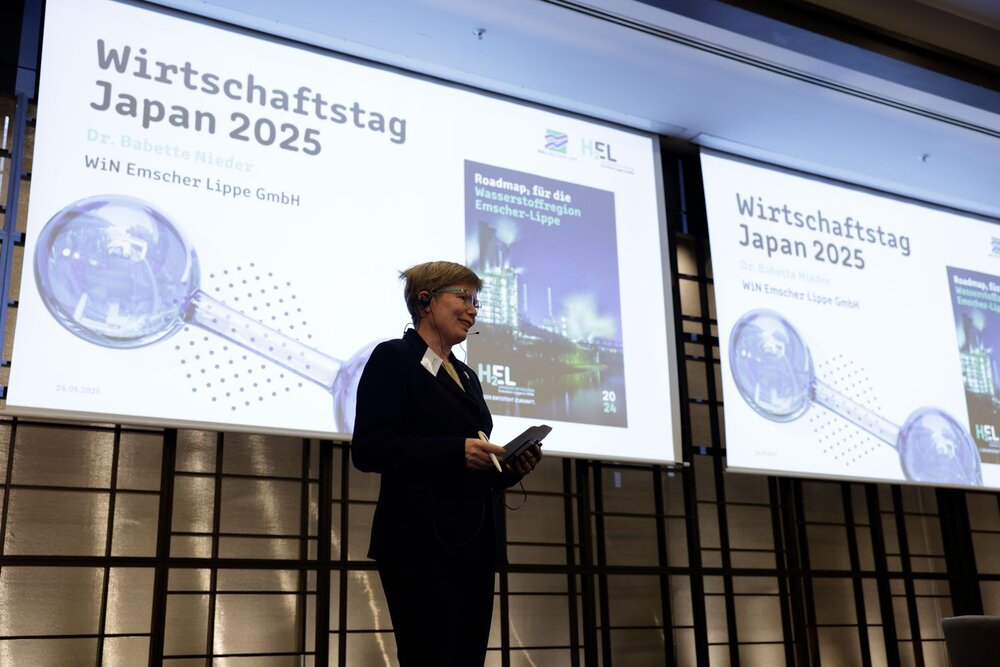 A woman in business attire wearing a headset stands on a stage, behind her projections with the words “Japan Business Day 2025”.