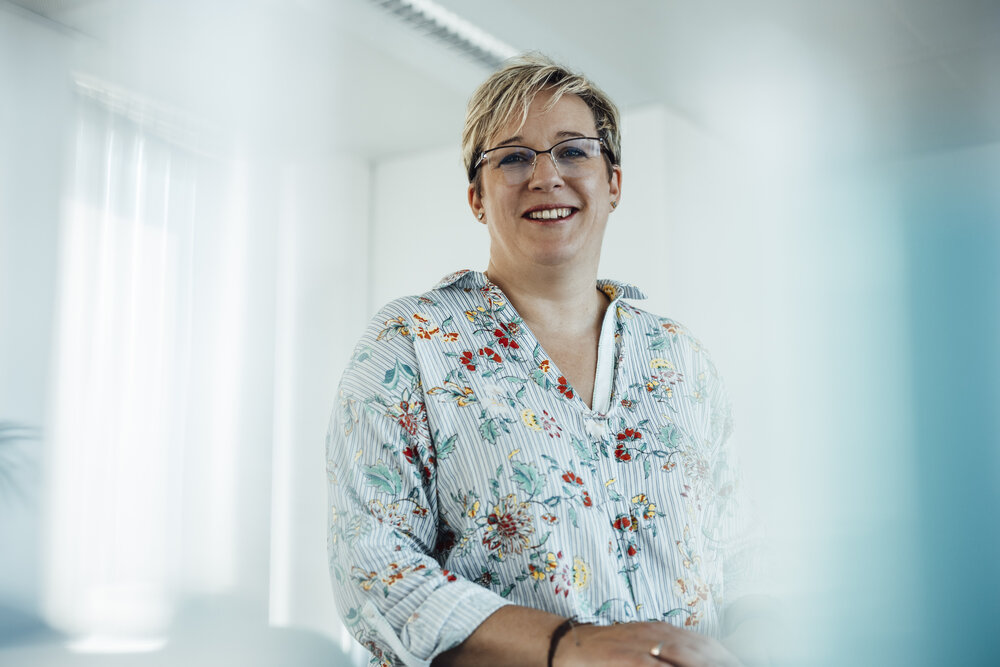 A portrait photograph of a woman with short brown-blonde hair. She is wearing glasses and a light-coloured patterned blouse. 