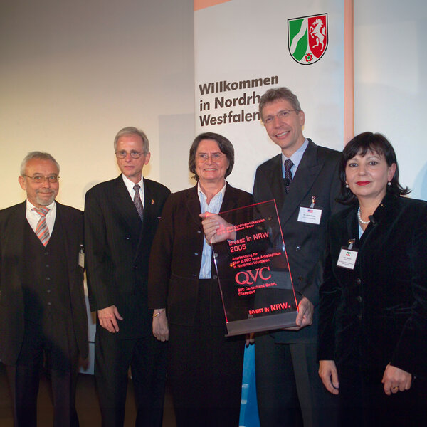 Five people in business attire stand in front of a banner displaying the coat of arms of North Rhine-Westphalia. A woman and a man in the centre hold an award.