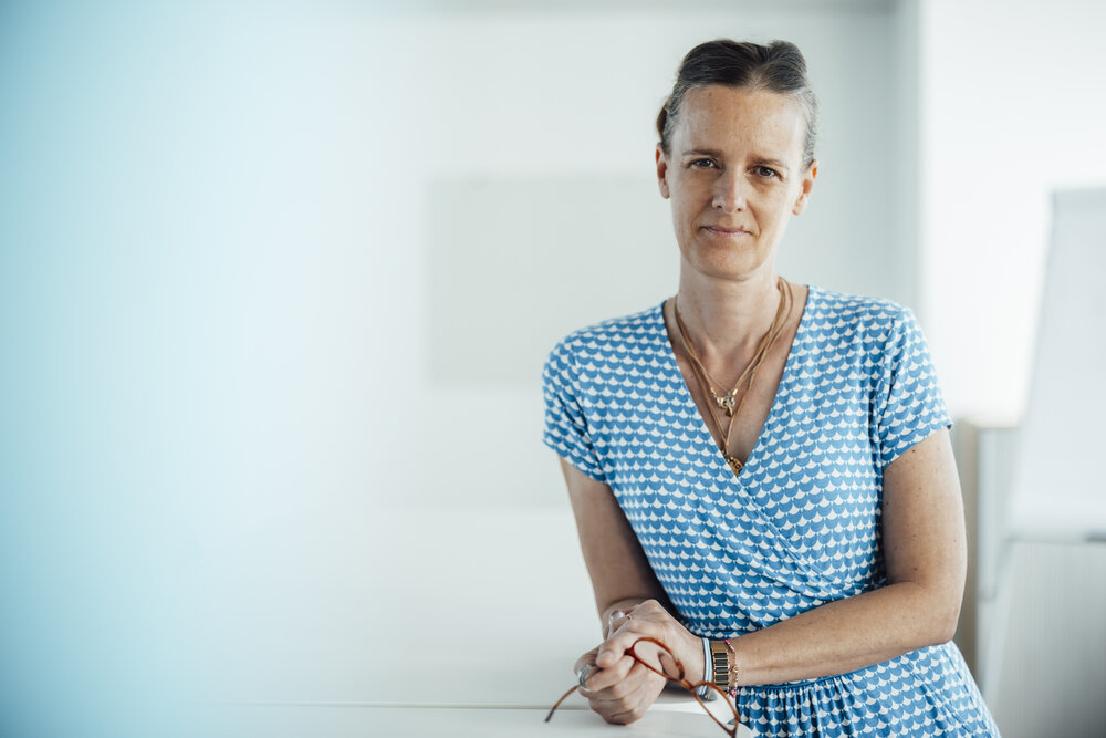 A portrait photo of a woman in a blue and white summer dress. She is standing at an office desk and holding her glasses in her hand.