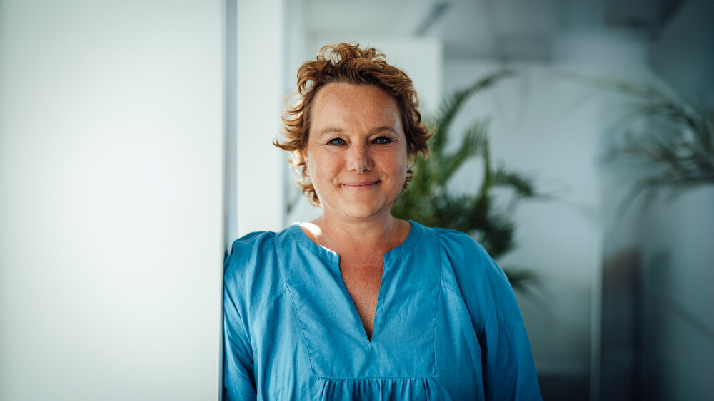 A portrait photograph of a woman wearing a blue blouse standing sideways against an office wall.