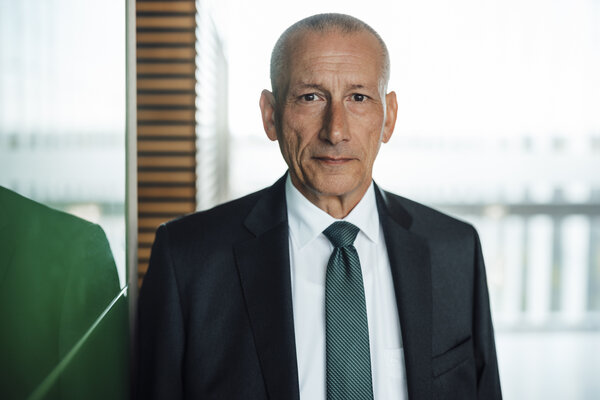 A portrait photo of a smartly dressed man. He is wearing a white shirt, a checked tie and a black jacket.