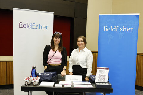 Two women stand behind a trade fair stand with brochures and giveaways, with a banner bearing the inscription “fieldfisher” in the background.