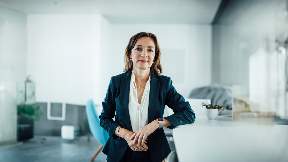 A portrait photo of a smartly dressed woman wearing various pieces of jewellery. She is standing sideways at an office desk with her arm resting on it.