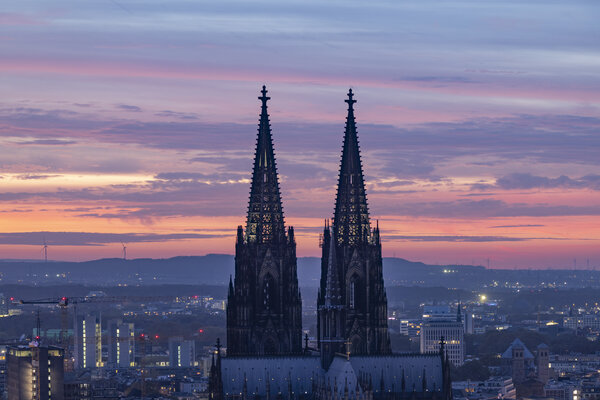 Die beiden Türme des Kölner Doms im Vordergrund bei Abenddämmerung, dahinter die Stadt Köln mit beleuchteten Gebäuden.