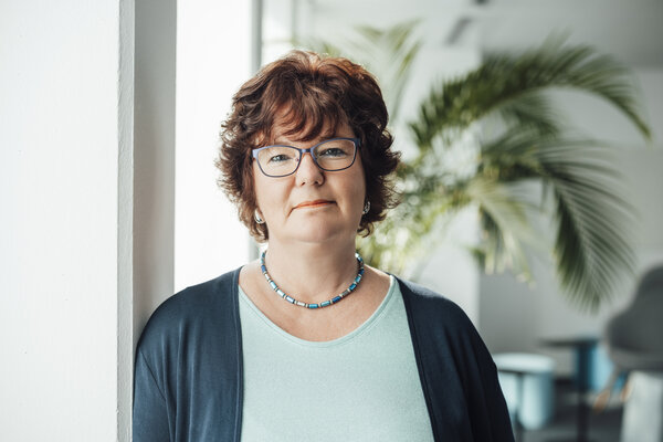 A portrait photograph of a woman. She is wearing glasses and a necklace, as well as clothing in various shades of green.