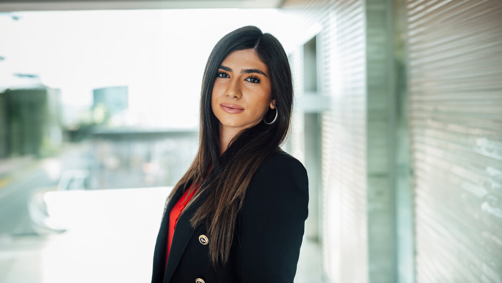 A portrait photograph of a woman wearing a black blazer. She is standing sideways to the camera.