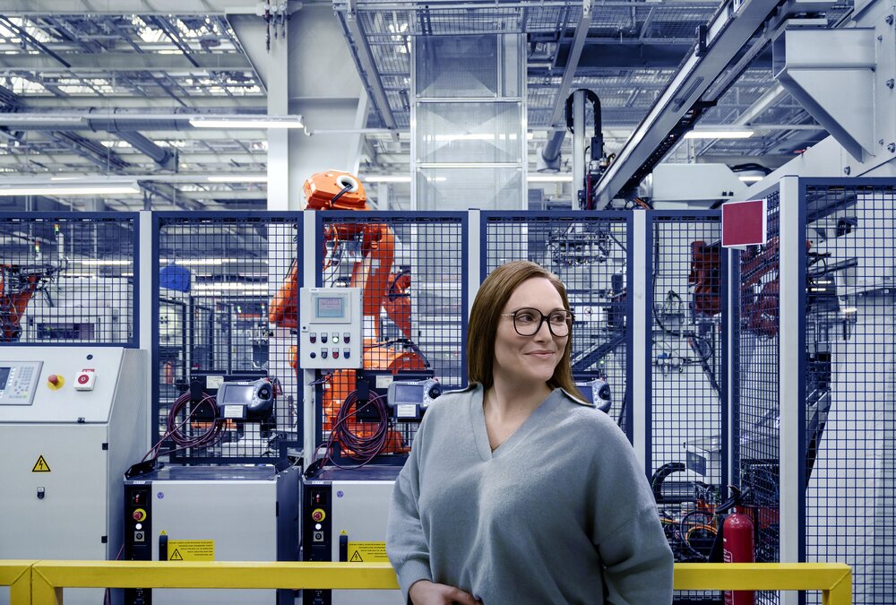 A woman wearing glasses stands in a modern factory hall in front of a cordoned-off area containing orange industrial robots.