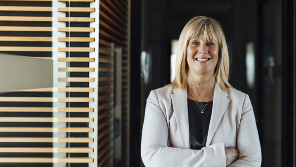 A portrait photograph of a smartly dressed woman. She has her arms crossed and is wearing a light grey blazer and a necklace.