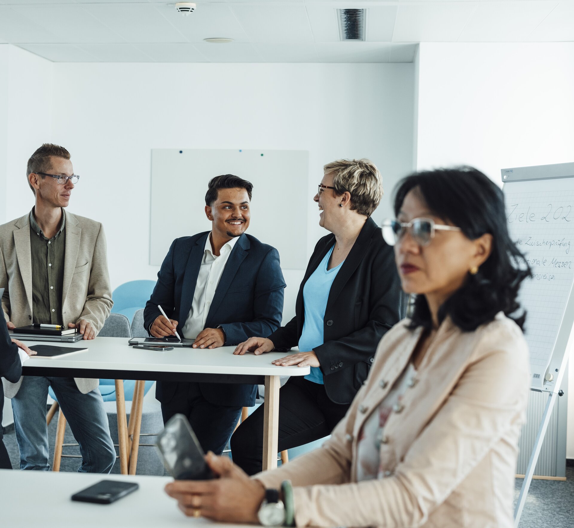 Several people in business attire are sitting and standing in a bright meeting room, with a flipchart containing notes in the background.
