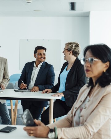 Several people in business attire are sitting and standing in a bright meeting room, with a flipchart containing notes in the background.
