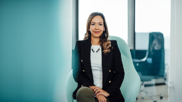 A portrait photograph of a woman sitting on a chair. She is wearing olive green trousers, a white shirt and a dark-coloured blazer.