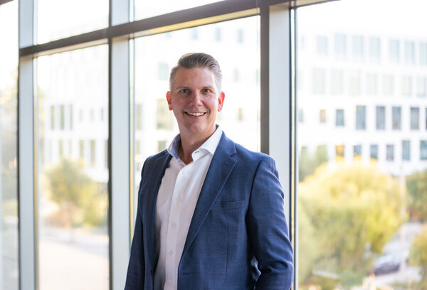 A portrait photograph of a smartly dressed man with styled hair standing in front of several windows.