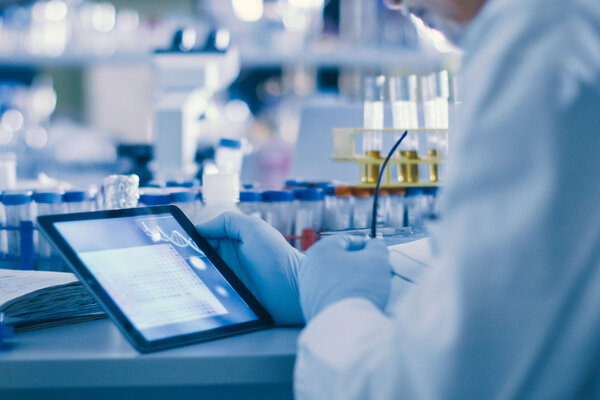 A person in a laboratory working on a tablet, surrounded by test tubes and notes.