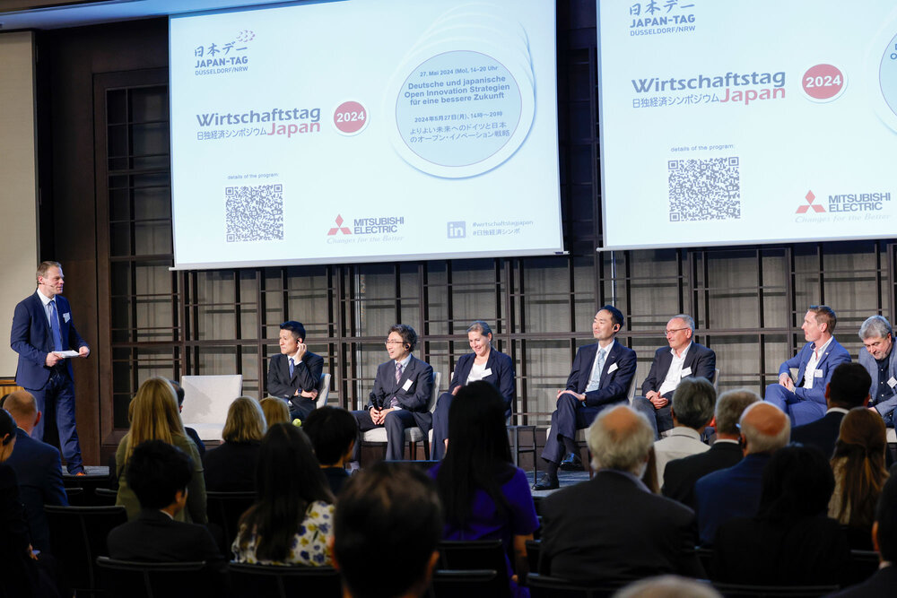 Several people in business attire are seated on chairs on a stage, with many more people seated in front of them.