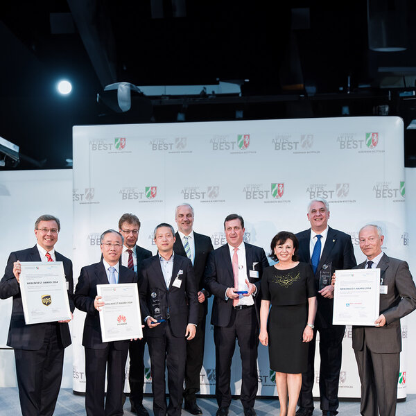 Nine people in business attire stand side by side, some holding certificates and glass trophies, with a screen in the background displaying the words “Germany at its best”.