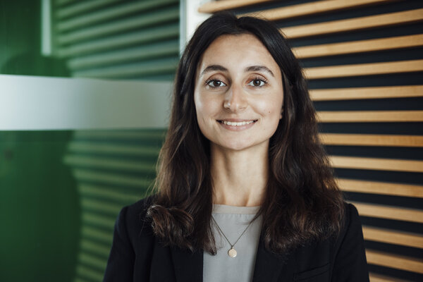 A portrait photograph of a young woman. She is wearing a silver necklace and standing next to a glass partition.