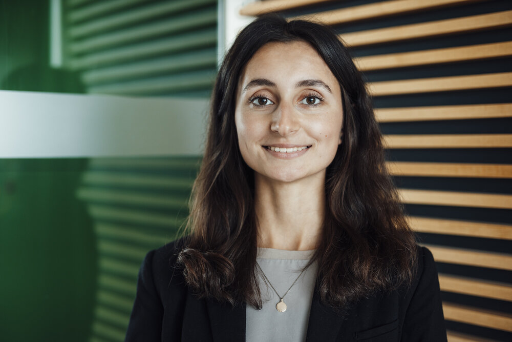 A portrait photograph of a young woman. She is wearing a silver necklace and standing next to a glass partition.