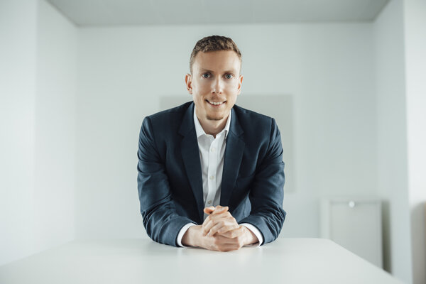 A portrait photograph of a smartly dressed man with styled blond hair sitting behind an office desk.